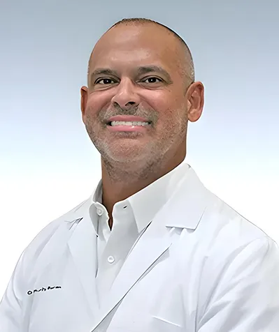 Dr. Rodolfo Alfonso smiling in a white lab coat, photographed against a light blue gradient background.